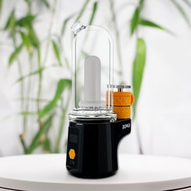 A sleek, black Bomb Sykloud E Rig with a glass cup and an orange heating chamber coating, shown on a white surface against a blurred background of green leaves.